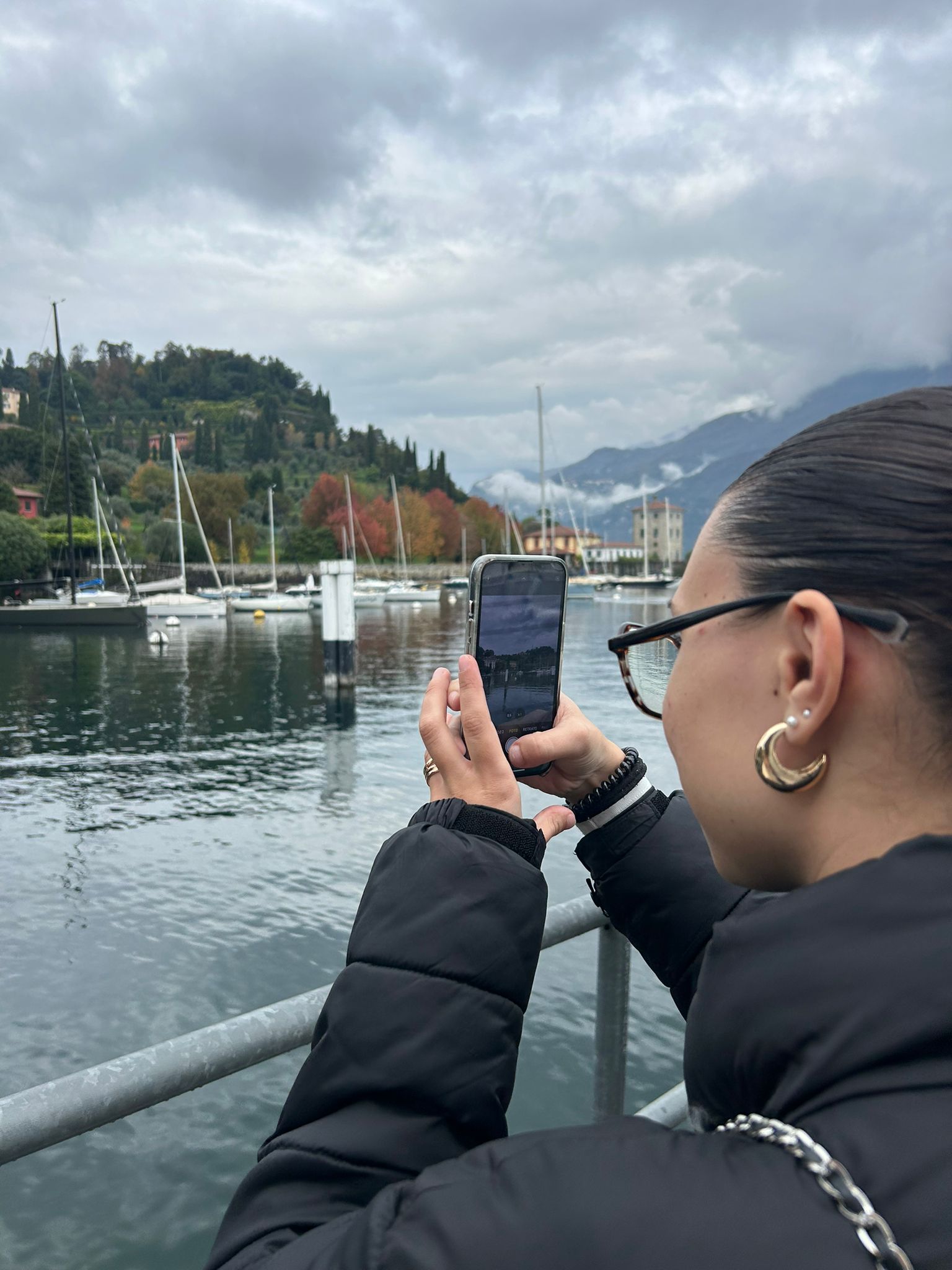 Woman taking a photo by a lakeside with sailboats and mountains in the background